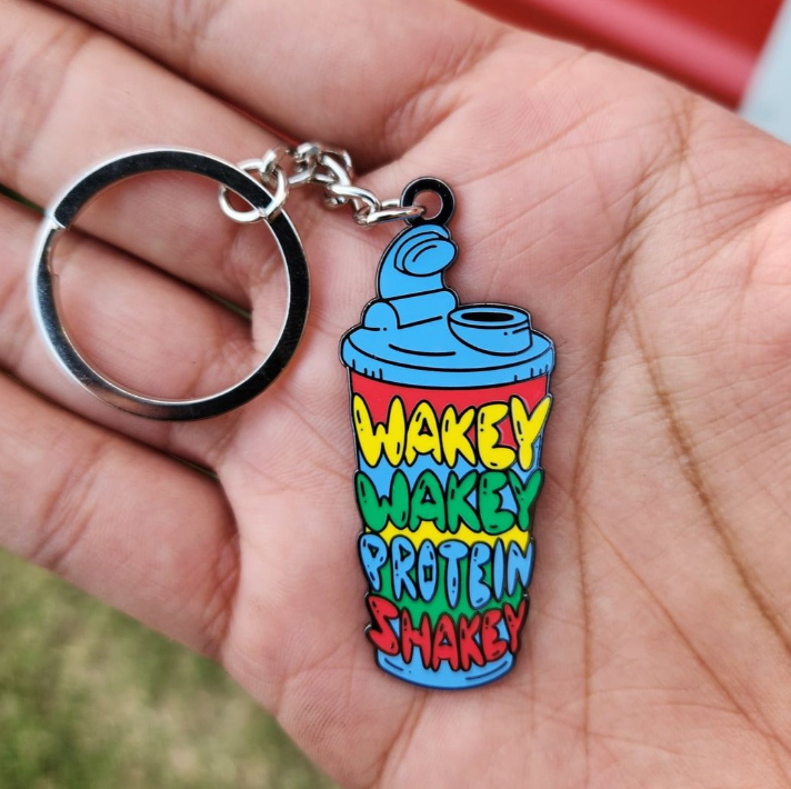 A hand holding a keychain with "WAKEY WAKEY PROTEIN SHAKEY" text